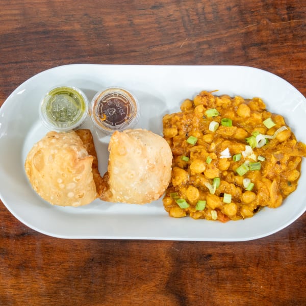 Two golden-fried samosas, chickpea curry, and two dipping sauces on a white plate.