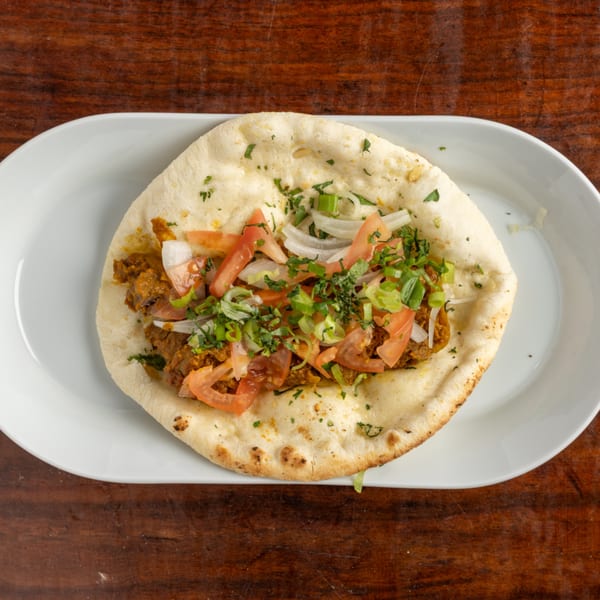 Naan bread topped with meat, sliced tomatoes, onions, and fresh herbs on a white plate.
