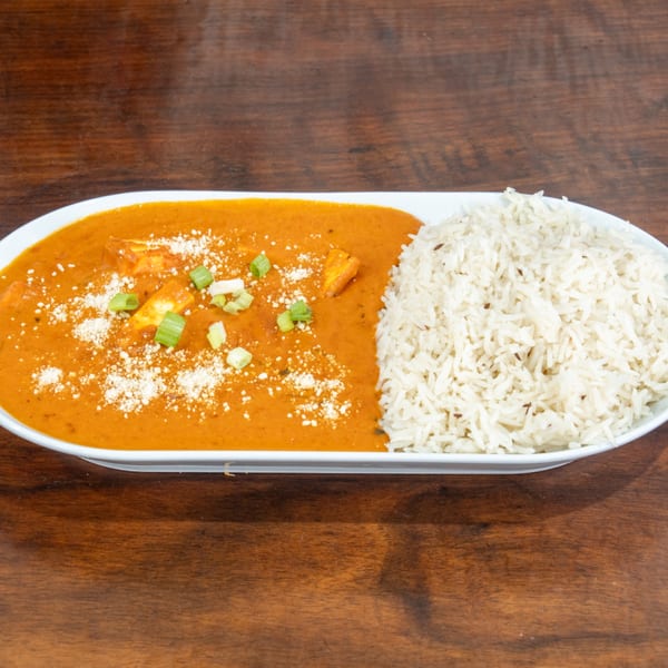 A white oval plate with orange curry, paneer, green onions, and white rice on a wooden table.