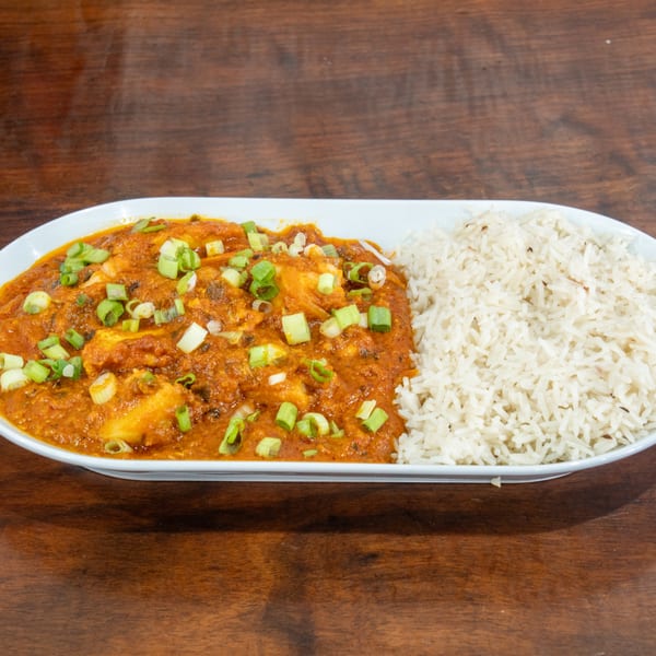 A white oval plate holds a vibrant orange curry with paneer and green onions, alongside white rice.